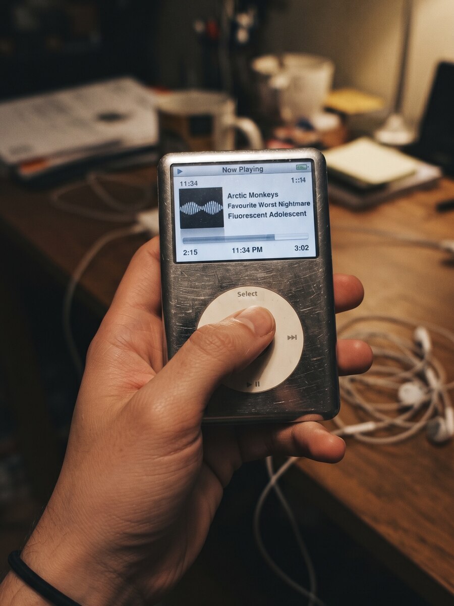 A scratched-up iPod Classic held in hand, screen showing Arctic Monkeys playing, earbuds tangled on the desk behind it