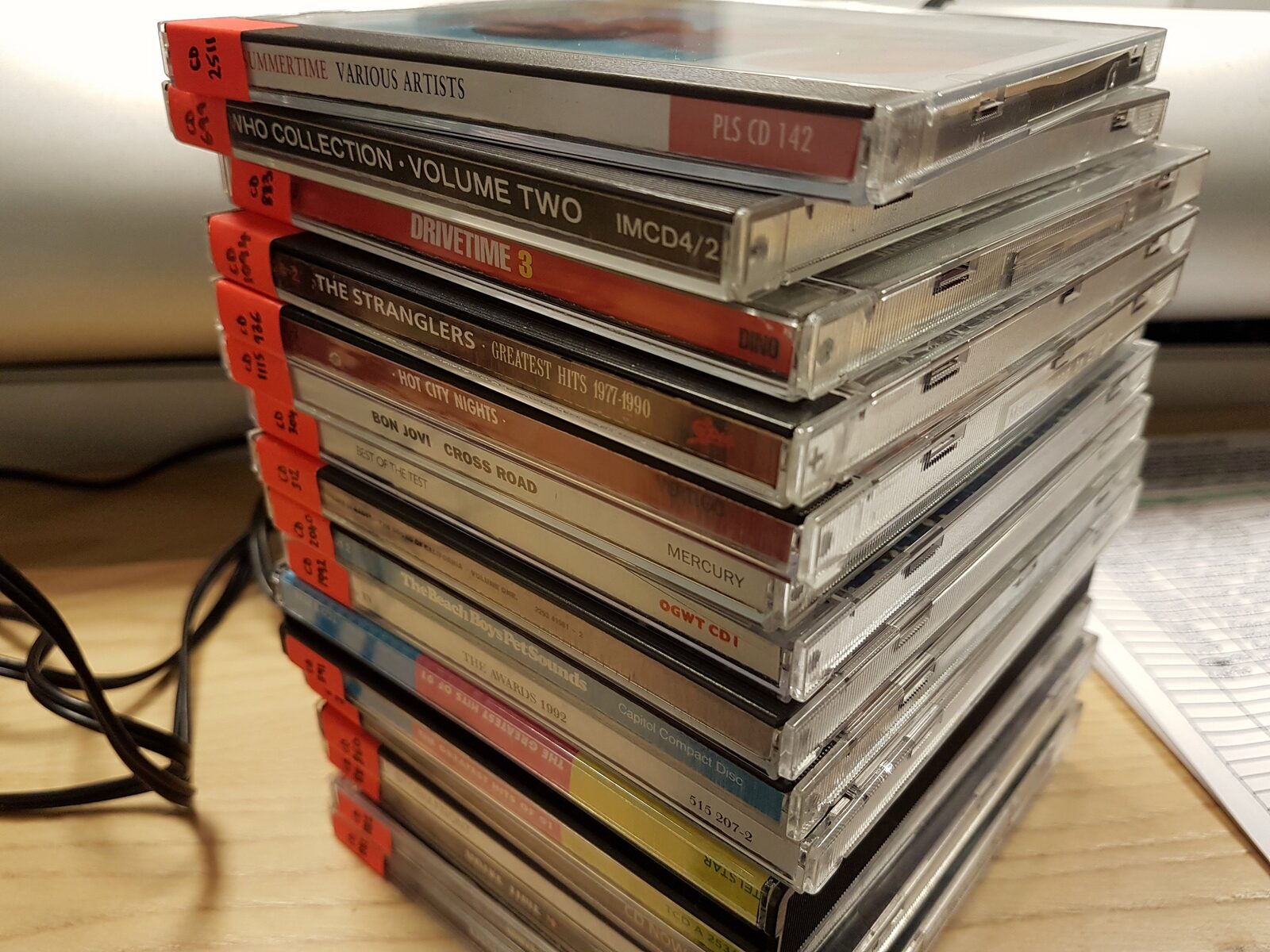 A stack of CDs in jewel cases on a desk, spines hand-labeled with red stickers